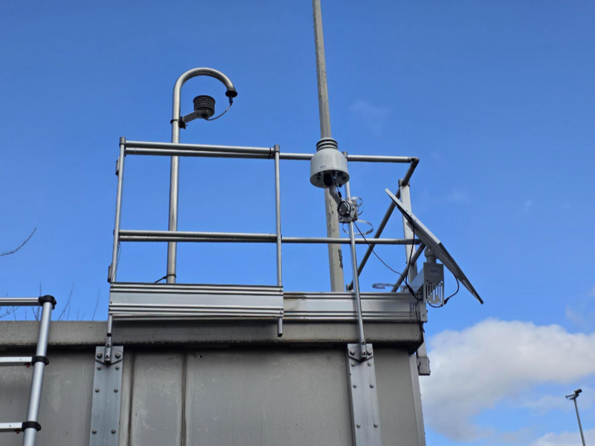 Outdoor sensor-based air quality monitor undergoing field comparison beside reference monitoring equipment.